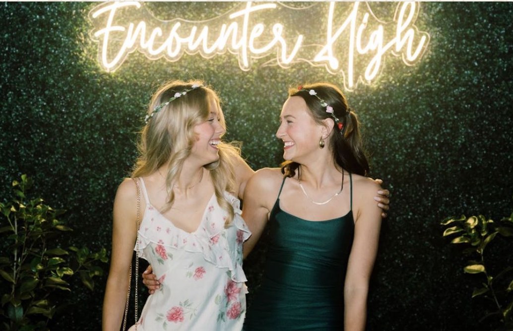 Two students laughing together beneath a glowing Encounter High neon sign