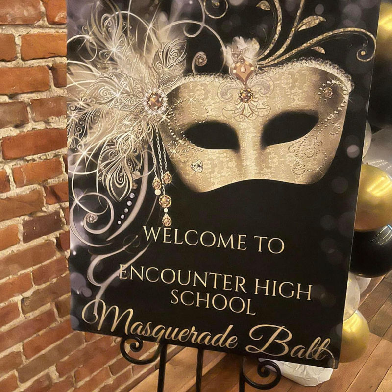 Welcome to Encounter High School Masquerade Ball sign beside gold and black balloons